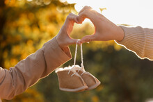 Man And Pregnant Wife Are Doing Heart Gesture With Hands And Holding Warm Baby Shoes On Nature Autumn Background. Future Parents Waiting For The Baby. The Concept Of Mother's Day And Women's Day.