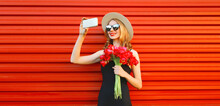 Portrait Of Beautiful Young Woman Taking Selfie With Smartphone Holding Bouquet Of Pink Flowers In Summer Straw Round Hat On Red Background