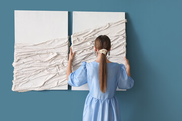 Wall Mural - Young woman hanging 3D picture on blue wall, back view