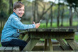 Happy young kid is sitting outside with a phone. Smiling teen boy outdoors in a park with a smartphone in his hands. The kid is distracted from the phone and looking at the camera.
