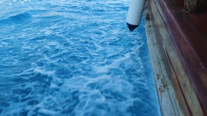 Wall Mural - Side view of a wooden boat sailing on the sea. In the background of the deep blue sea, the boat is broken up by glistening white foamy waves. Rear view. Slow motion