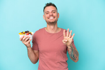 Wall Mural - Young caucasian man holding a bowl of fruit isolated on blue background happy and counting four with fingers