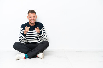 Wall Mural - Young man sitting on the floor isolated on white background pointing to the front and smiling