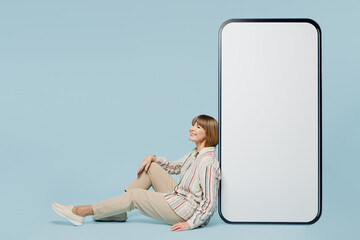 Poster - Full body side view elderly woman 50s year old in shirt sitting near big huge blank screen mobile cell phone smartphone with area isolated on plain pastel light blue color background studio portrait