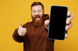 © ViDi Studio - Young redhead man wear brown hoody casual clothes hold in hand use close up mobile cell phone with blank screen workspace area show thumb up wink isolated on plain yellow background studio portrait.