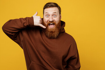 Young overjoyed excited fun redhead caucasian man wearing brown hoody casual clothes doing phone gesture like says call me back isolated on plain yellow background studio portrait. Lifestyle concept.