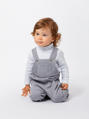 A Caucasian toddler of 2 years old with curly hair in a gray jumpsuit and a white turtleneck is kneeling and looking sadly away.