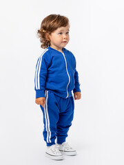 A cute curly-haired toddler of 1-2 years old stands sideways in a blue tracksuit and sneakers on a white one and looks away.