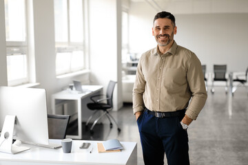 Successful person. Portrait of happy middle aged businessman posing with hands in pockets and smiling, office interior