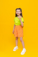 Wall Mural - A child in shorts and a T-shirt. Happy childhood. A little girl in full height with long hair rejoices in the summer warmth inside and points to your advertisement on a yellow isolated background.