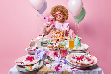 Canvas Print - Positive birthday woman holds cellphone in front of herself and toasts dressed in festive outfit poses near table with piles of desserts surrounded by inflated balloons isolated over pink background