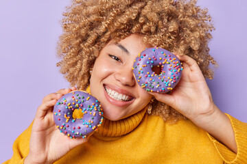 Wall Mural - Happy sincere curly haired European woman covers glazed doughnuts over eye smiles broadly being in good mood enjoys eating harmful sweet food wears yellow jumper isolated on purple background