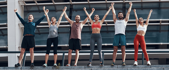 group of happy young people in sports clothing keeping arms raised while standing outdoors