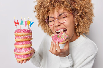 Wall Mural - Hungry woman has sweet tooth eat delicious glazed doughnut celebrates birthday alone wears spectacles and jumper enjoys eating favorite dessert isolated over white background. Celebration concept