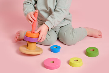 Wall Mural - Happy toddler baby plays with a pyramid on a studio pink background. Child boy puts wooden rings on a toy pyramid. Kid age one year eight months