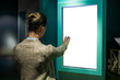Woman using white blank interactive touchless vertical display of electronic multimedia kiosk with no touch control in dark room. Mock up, copyspace, template and technology concept