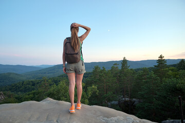 Wall Mural - Sportive woman standing alone on hillside trail. Female hiker enjoying view of evening nature from rocky cliff on wilderness path. Active lifestyle concept