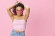 © SHOTPRIME STUDIO - Happy woman hands up waxing her armpits, shaving, with curly hair in a pink tank top and jeans on a pink background wearing sunglasses with a nice tan, copy space