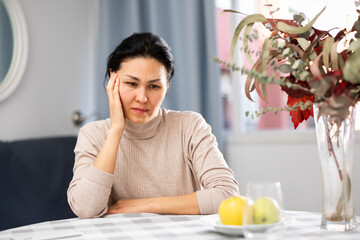 Upset Asian woman sitting at table at home. Portrait of sad woman.