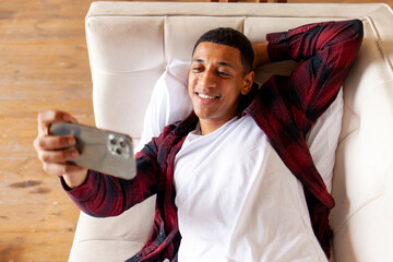 Wall Mural - young african american man lies on comfortable sofa and takes selfie with smartphone, guy with phone communicates