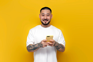 Wall Mural - adult tattooed asian man in white t-shirt uses smartphone on yellow isolated background and smiles