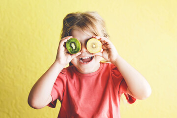 Wall Mural - Happy little child girl with yellow and green kiwi fruits on yellow background. Preschool girl smiling. Healthy fruits for children