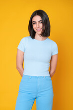 Smiling Young Woman With Hands Behind Back Standing Against Yellow Background