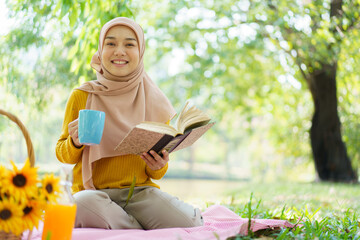 Wall Mural - Happy cheerful Asian beautiful muslim woman sitting on the ground at the park and drinking a cup of tea in afternoon. Beautiful muslim woman relaxing and picnic in weekend.