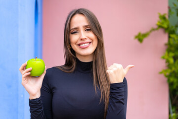 Wall Mural - Young pretty woman with an apple at outdoors pointing to the side to present a product