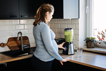 Wall Mural - Green Smoothies Recipes For Pregnancy and Postpartum, Prenatal Nutrition. Pregnant woman preparing green vitamin smoothie with a blender and drinking in the kitchen.