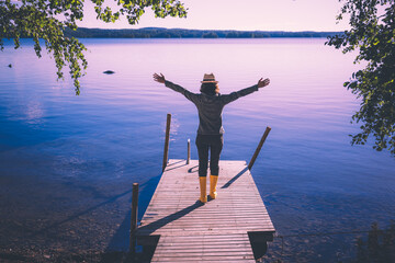 Girl enjoying nature looking at beautiful blue lake. Woman stands with raised arms on wooden pier in Finland.