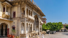 The Facade Of The Ancient City Palace In Jaipur.  The Building With Columns, Arches, Balconies Is Decorated With Carvings. There Are Sculptures Of Elephants At The Entrance. Blue Sky. India.
