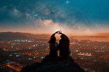 Silhouette Rear View Of A Heterosexual Couple Sitting On Top Of A Mountain Making A Silhouetted Heart Shape Sign With Their Hands Against The Background Of Moon And Stars Over The City