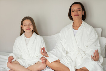 Wall Mural - Smiling young woman and little caucasian girl in bathrobe with closed eyes, meditation, resting on white bed