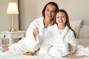 Wall Mural - Happy european millennial woman and little girl sit on bed, enjoy vacation together, weekend and cup of drink