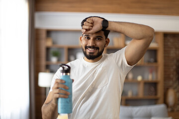 Wall Mural - Tired indian man in sport clothes wiping sweat and drinking water during morning exercise. Active young guy having regular workout during free time at home.