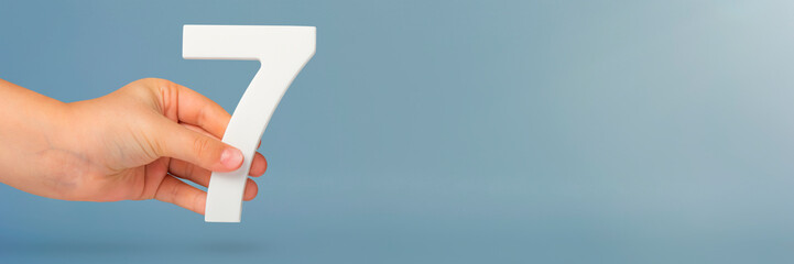 Fototapete - Number seven in hand. Hand holding white number 7 on blue background with copy space. Concept with number seven. Birthday 7 years, percent, seventh grade or day.