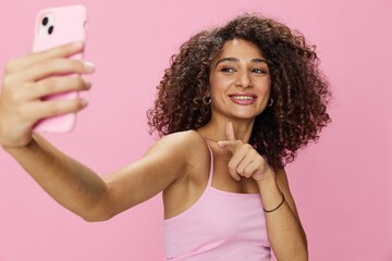 Woman blogger holding phone video call takes selfies, with curly hair in pink smile t-shirt and jeans poses on pink background, copy space, technology and social media, online