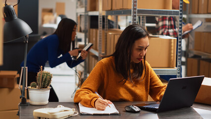 Young woman organizing merchandise in piles, putting retail products in packages for distribution. Female entrepreneur using laptop to check inventory and work on financial planning.