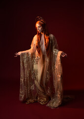 Wall Mural - Full length fantasy portrait of beautiful woman model with red hair, goddess silk robes & gold crown. Standing pose gestural hands reaching out isolated on dark red studio background 