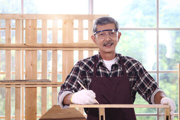 Portrait of happy smiling senior Asian carpenter wearing safety glasses, hold pencil in woodwork carpentry, old citizen craftsman working in joinery workshop, cheerful elderly man enjoy DIY hobby