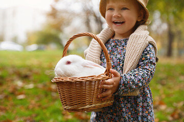 Wall Mural - Happy girl holding basket with cute white rabbit in autumn park, focus on pet