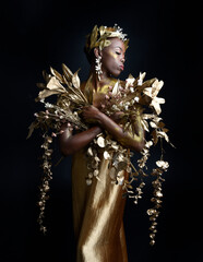 Wall Mural -  fantasy portrait of beautiful african woman model with afro, goddess silk robes and ornate floral wreath crown. gestural Posing holding golden flowers. isolated on dark  studio background 