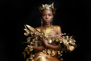 Wall Mural -  fantasy portrait of beautiful african woman model with afro, goddess silk robes and ornate floral wreath crown. gestural Posing holding golden flowers. isolated on dark  studio background 