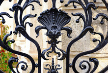 Black Wrought Iron Fence Detail In Budapest, Castle District. Forged Steel Decorative Elements. Ornate Sea Shell Shape. Steel Bar Grille. Soft Blurred Stone Building Elevation Background