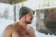 The sweaty man in a hat after sauna outdoors in winter. Steam due to temperature difference. Selective focus	
