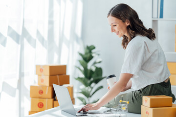 Delivery business. happy modern female in White Shirt checking order with laptop and holding a cup of coffee parcels using smartphone applications in the warehouse.