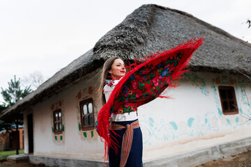 Wall Mural - Charming woman with traditional ukrainian handkerchief, necklace and embroidered dress standing at background of decorated hut with thatched roof. Ukraine, style, folk, ethnic culture