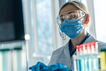 Wall Mural - A female researcher sits at a workplace in a laboratory, behind a personal computer monitor. Against the background research statistics. Pharmaceutical medical worker in protection works at keyboard.