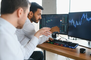 Two successful trader sitting in office and looking at screen, checking cryptocurrency information data on finance market graph, pointing on monitor